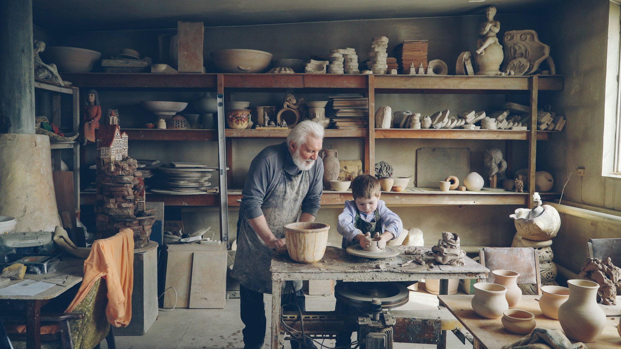 Grandfather and grandchild working clay at a table in a small pottery workshop lined with shelves